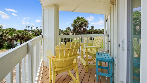 A wooden deck with yellow and blue Adirondack chairs, a small table, and a view of palm trees and a beach.