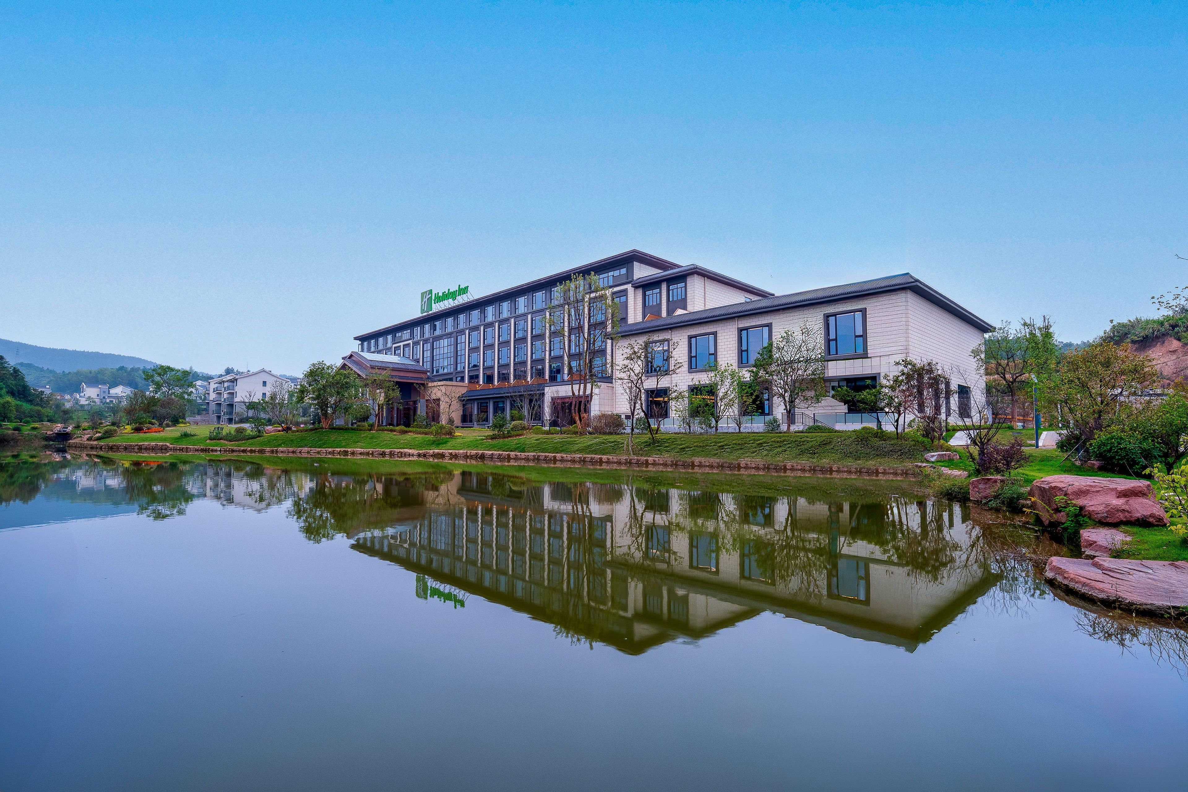 Ein modernes Hotel mit Spiegelung im Wasser, umgeben von viel Grün und blauem Himmel.