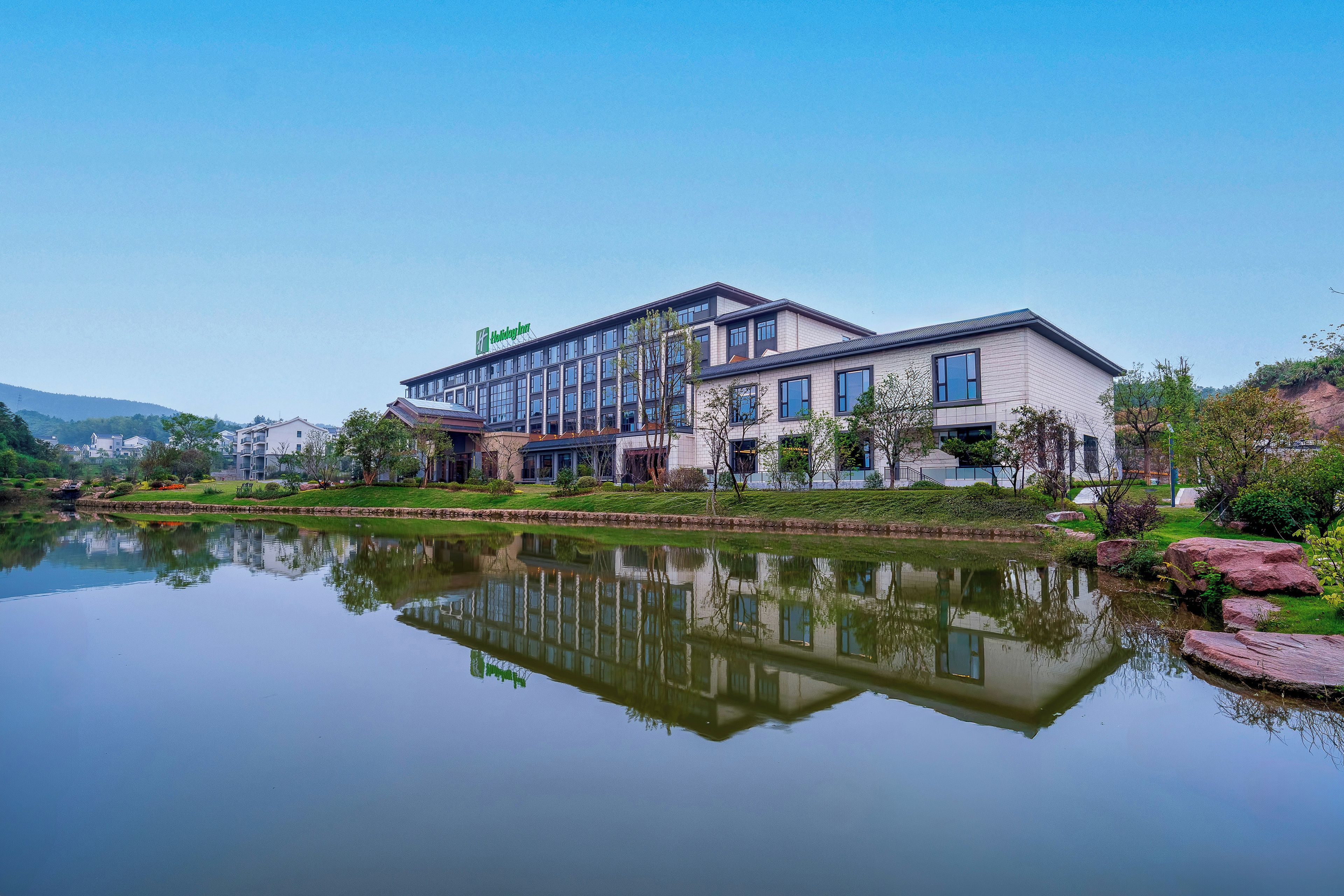 A Holiday Inn hotel with a reflection in the water, surrounded by greenery and a clear sky.