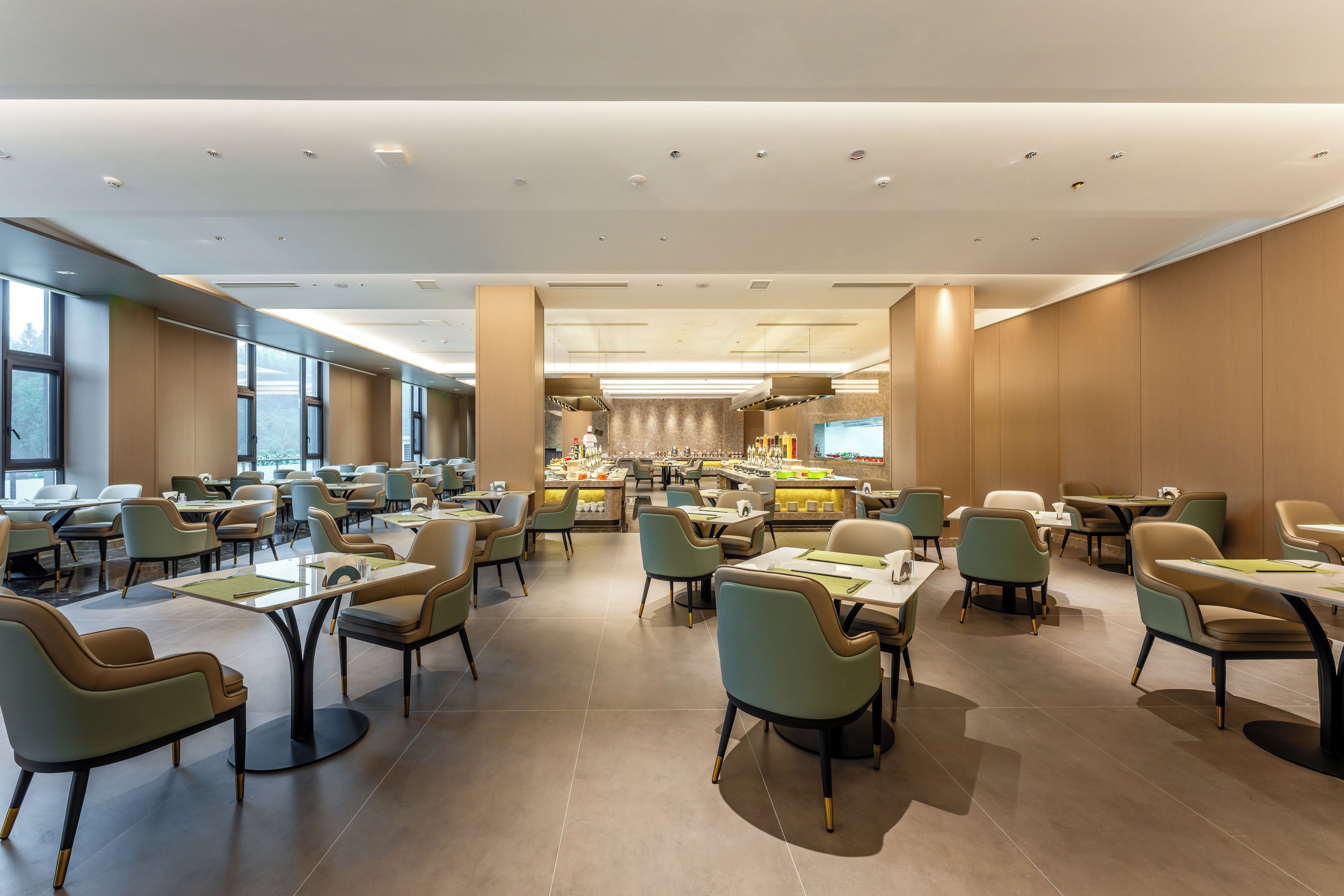 A spacious hotel dining area with multiple tables and chairs, a central bar, and large windows providing natural light.