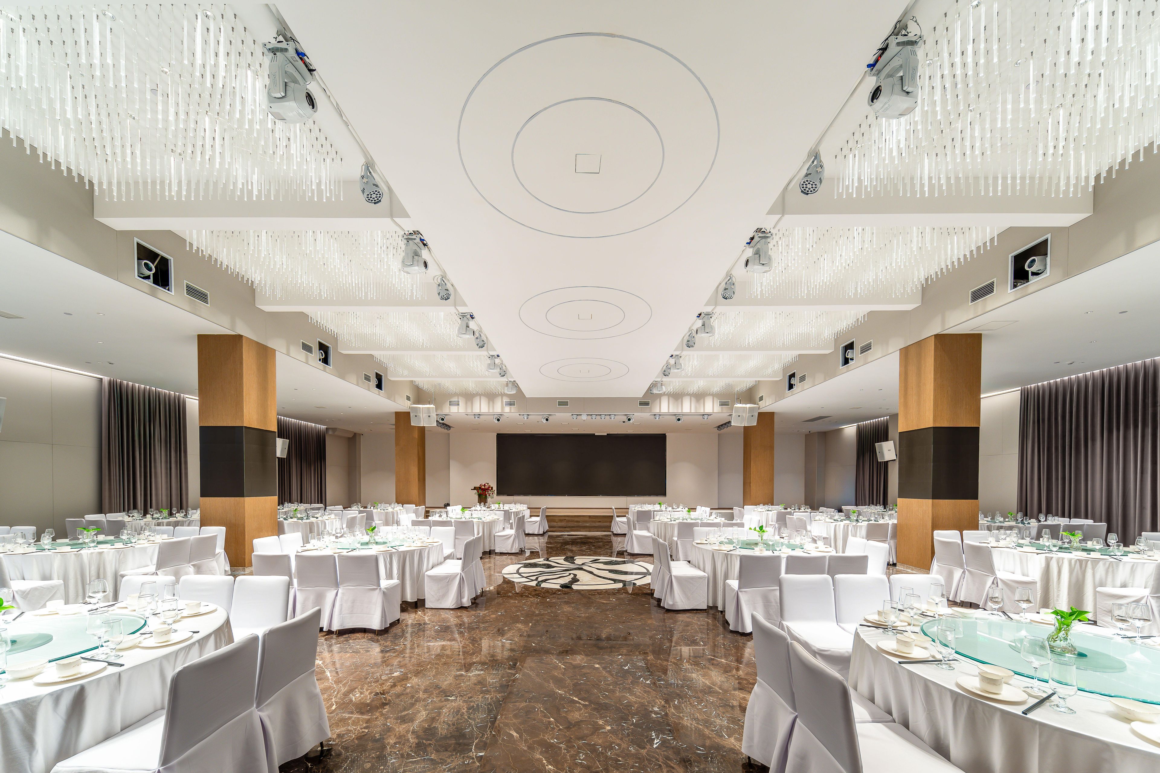 Un amplio salón de banquetes con mesas redondas, sillas blancas y un escenario central.