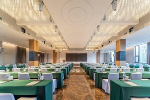 Un salón de conferencias con filas de mesas, sillas y una pantalla grande al frente.