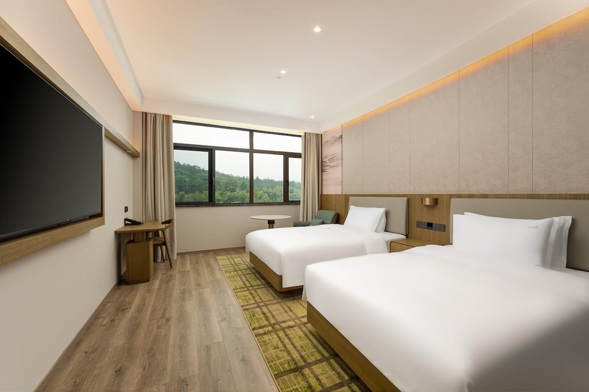 A hotel room with two beds, a desk, a chair, a small table, and a large window offering a view of greenery.