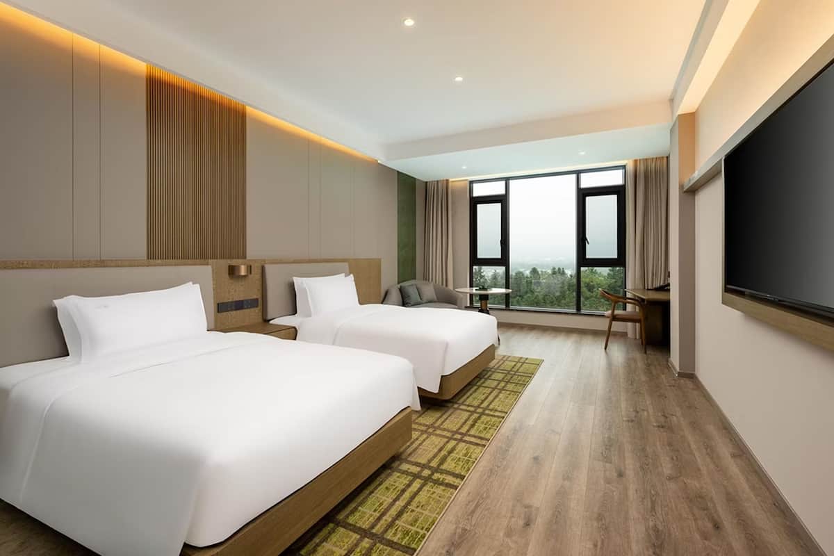 A hotel room with two beds, a desk, a chair, and a large window offering a view.