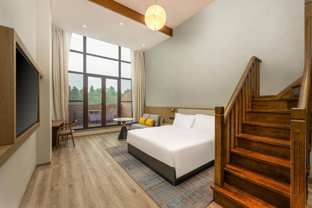 A hotel room with a large bed, wooden staircase, and a view of greenery through the window.