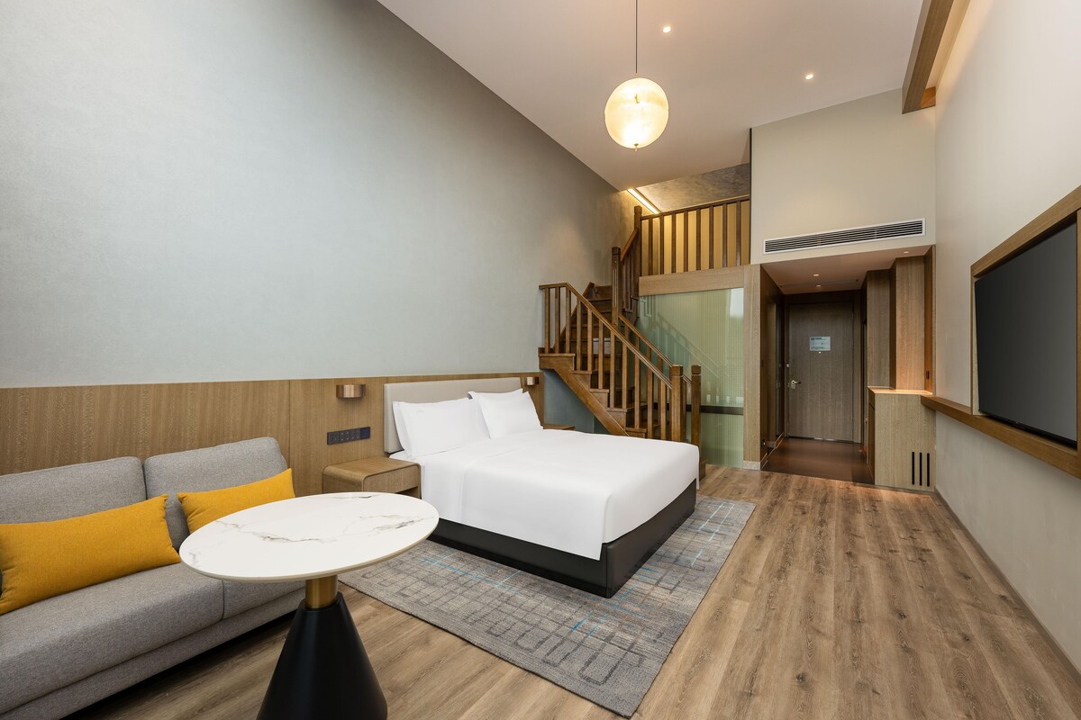 A modern hotel room with a bed, a sofa, a small table, and a staircase leading to an upper level.