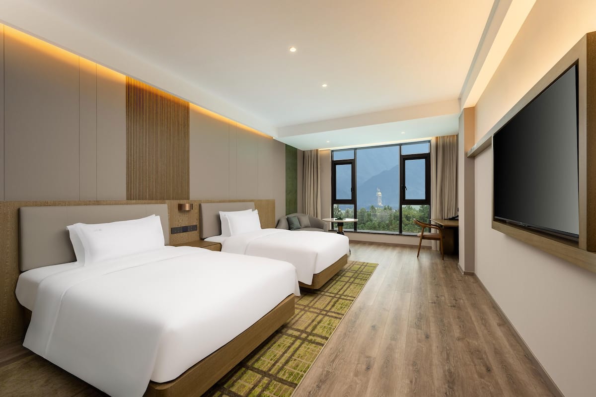 A hotel room with two beds, a desk, and a large window offering a mountain view.