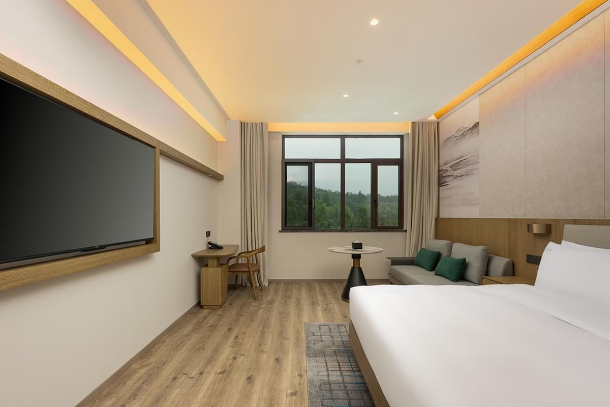 A modern hotel room with a large bed, a desk with a chair, a small table, and a large window offering a view of the outdoors.