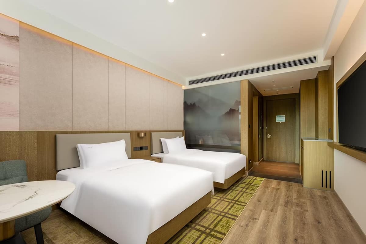 A hotel room with two beds, a small table, and a large window.