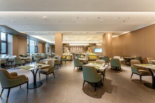 Un amplio comedor de hotel con numerosas mesas y sillas, una zona de bar central y ventanas grandes que proporcionan luz natural.