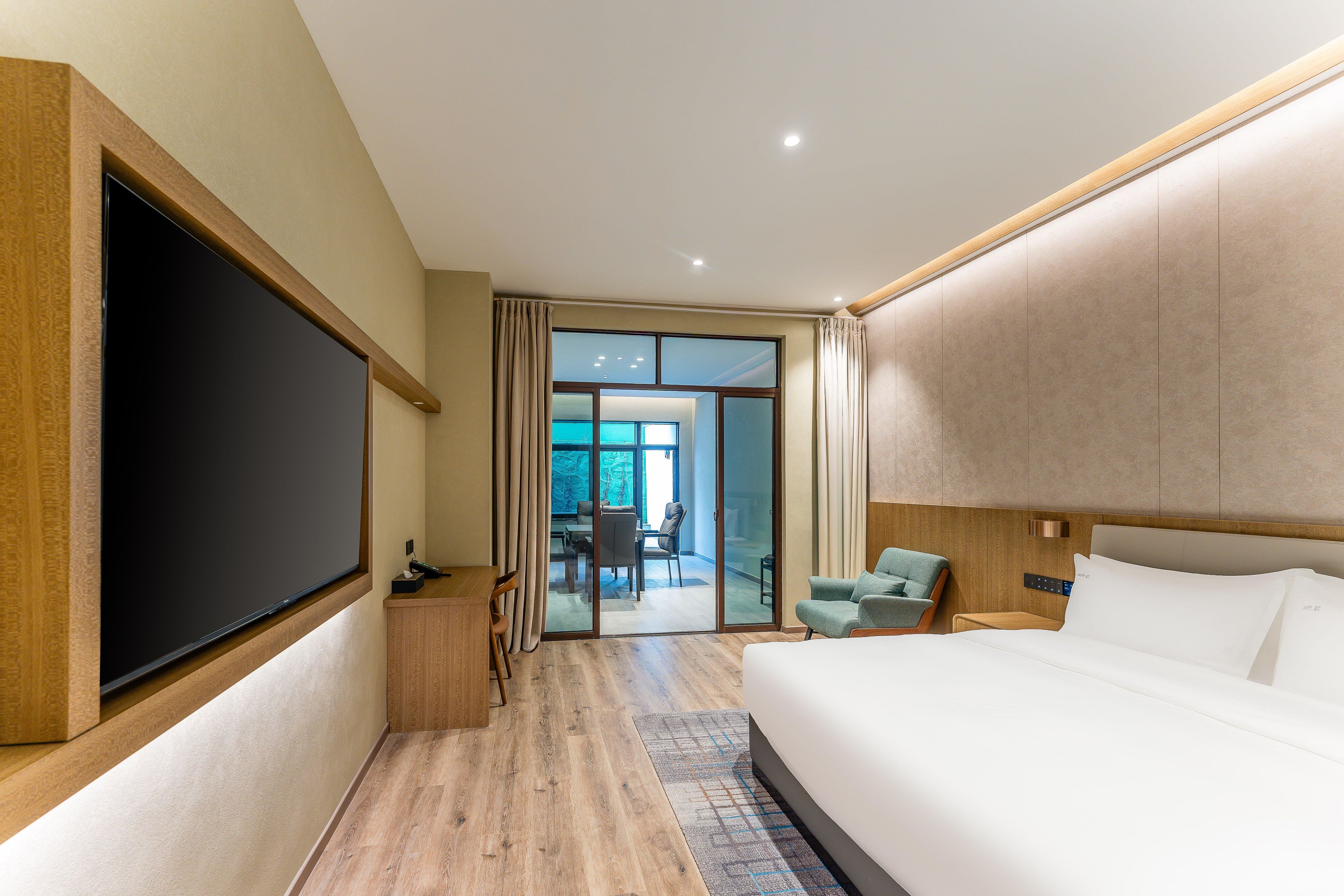 A modern hotel room with a large bed, a flat-screen TV, a desk with a chair, and a view of the outside through a sliding glass door.