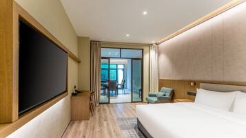 A modern hotel room with a large bed, a flat-screen TV, a desk with a chair, and a view of the outside through a sliding glass door.