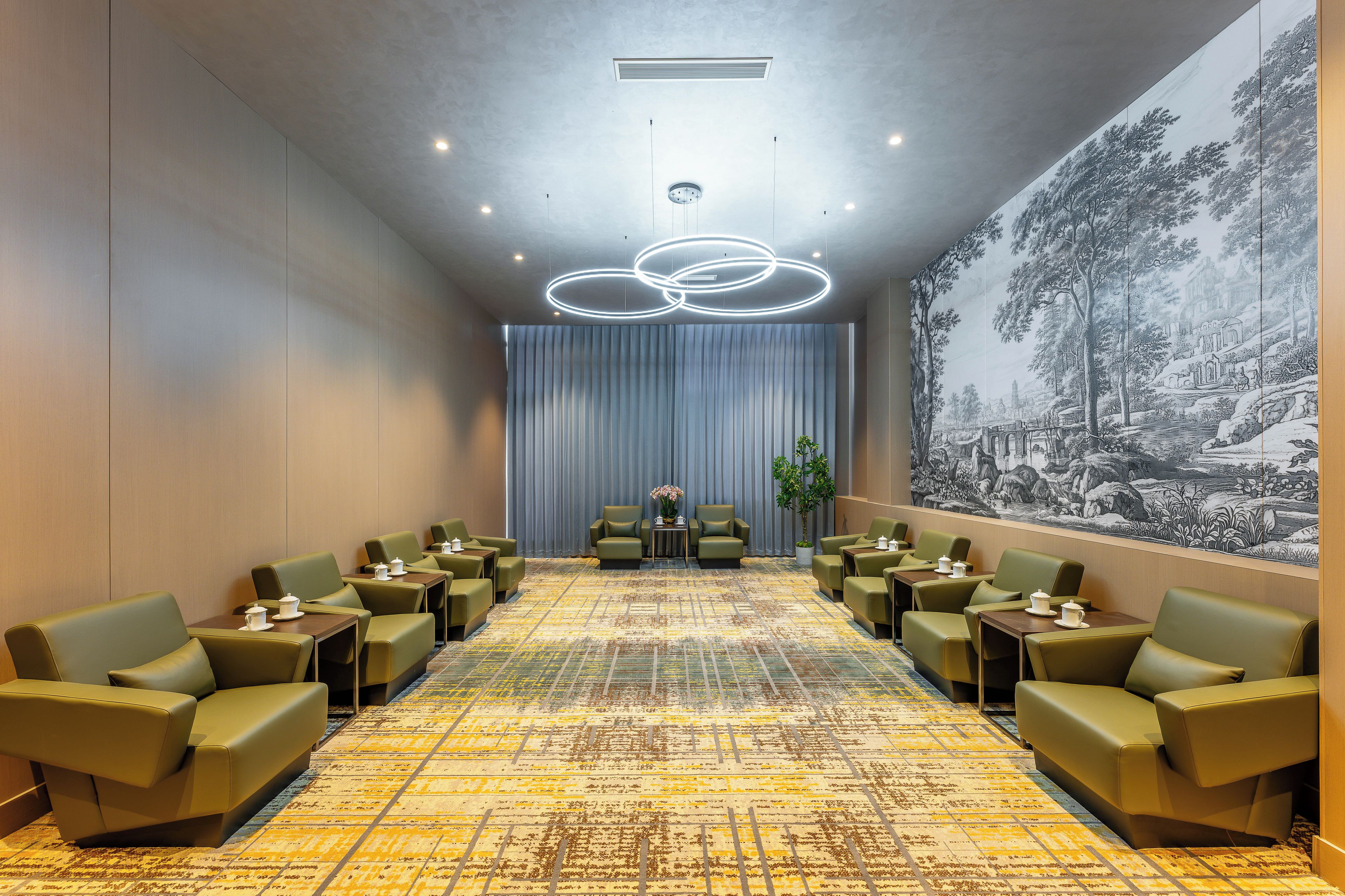 Een hotel lobby met een kamerbreed tapijt met patroon, groene zitplaatsen, een groot muurschilderij en een plafondlamp.
