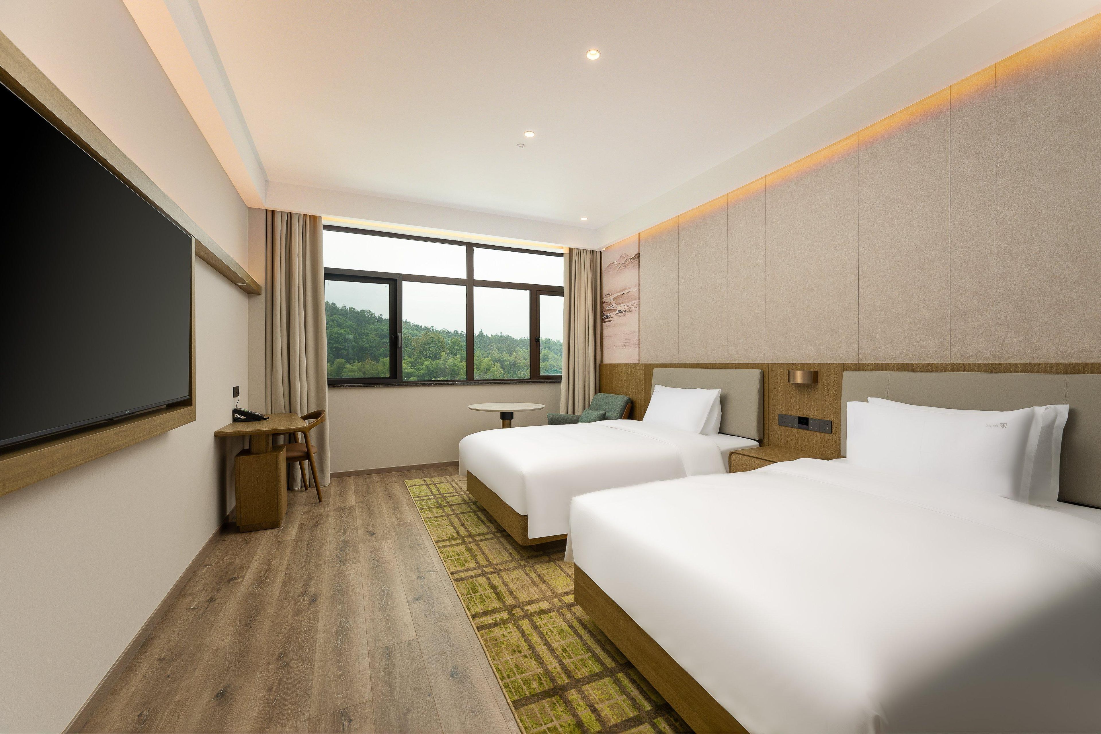 A hotel room with two beds, a desk, a chair, a small table, and a large window offering a view of greenery.