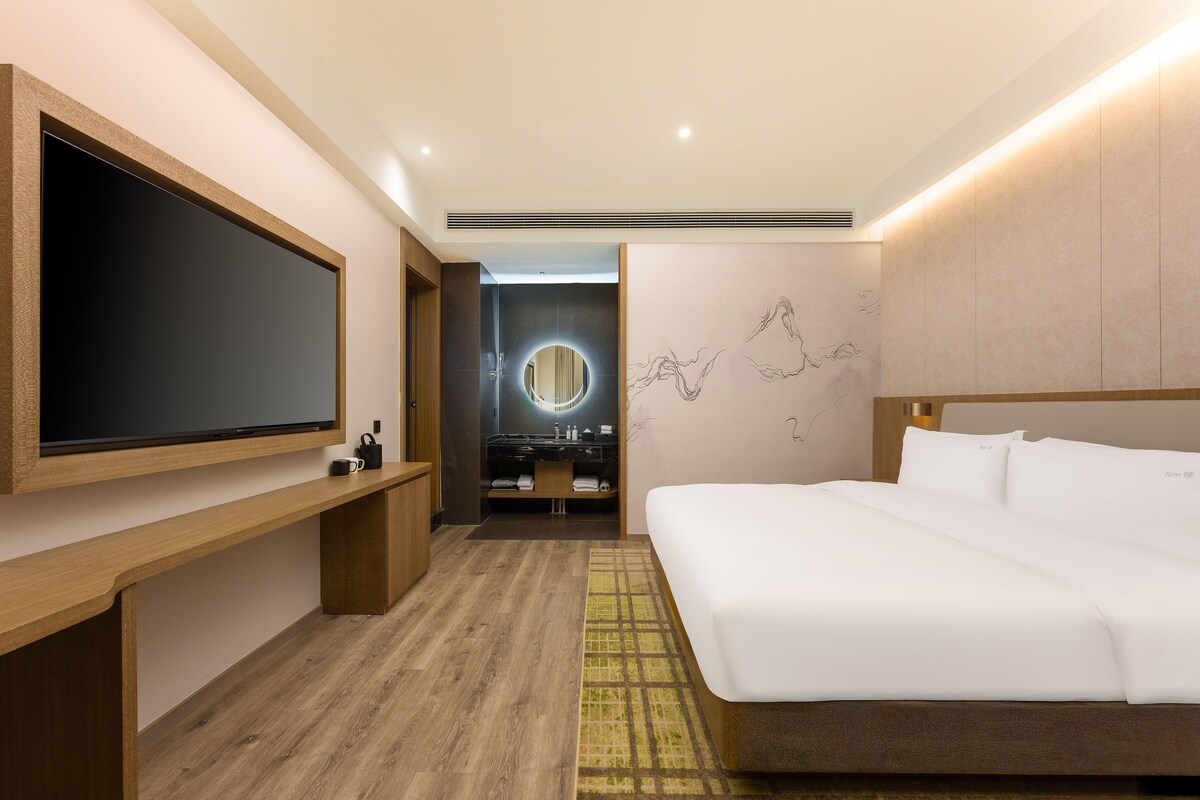 Quarto de hotel moderno com uma televisão de tela grande, um banco de madeira, uma cama com lençóis brancos, um espelho redondo e uma parede com arte abstrata.