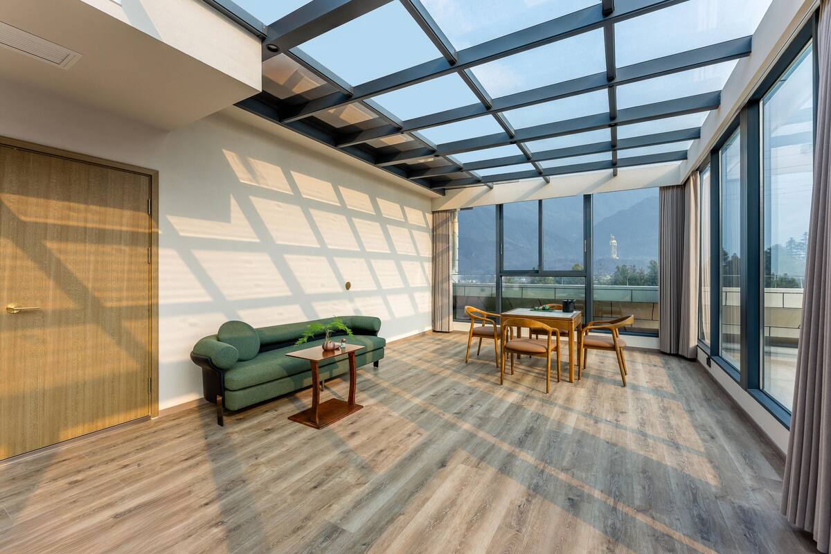 A spacious room with a high ceiling, large windows, a green sofa, a wooden table, and a view of mountains.