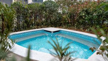 Una piscina rectangular con borde de azulejos, rodeada de abundante vegetación y un edificio al fondo.