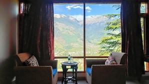 Une pièce dotée d’une grande fenêtre donnant sur des montagnes enneigées et un village situé en contrebas. On y trouve deux fauteuils en osier et une petite table sur laquelle sont posés une bouteille et des verres.