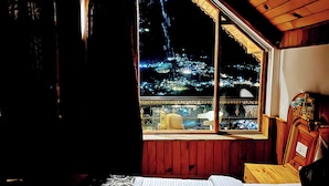 Une pièce dotée d’une grande fenêtre donnant sur une vue nocturne de la ville, d’un lit recouvert d’une couvre-lit à motifs et d’un plafond en bois.