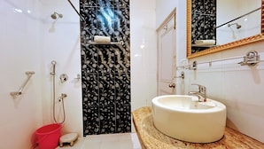 Une salle de bain avec un lavabo blanc, un miroir, une douche dont le mur est recouvert de carreaux noirs à motifs floraux et un seau rouge.