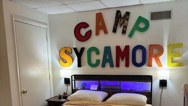 Un dormitorio con una cama, una mesita de noche, una lámpara y una pared con las palabras "CAMP SYCAMORE" en letras coloridas.