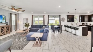 Una sala moderna con sofá, mesa de centro y televisor, un comedor con sillas y mesa, y una cocina con armarios oscuros y una barra.