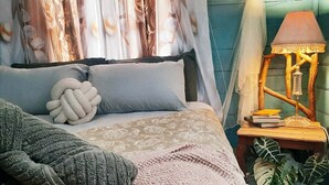 A bed with a patterned bedspread, a knitted throw, and a decorative pillow. There is a bedside table with a lamp and books.