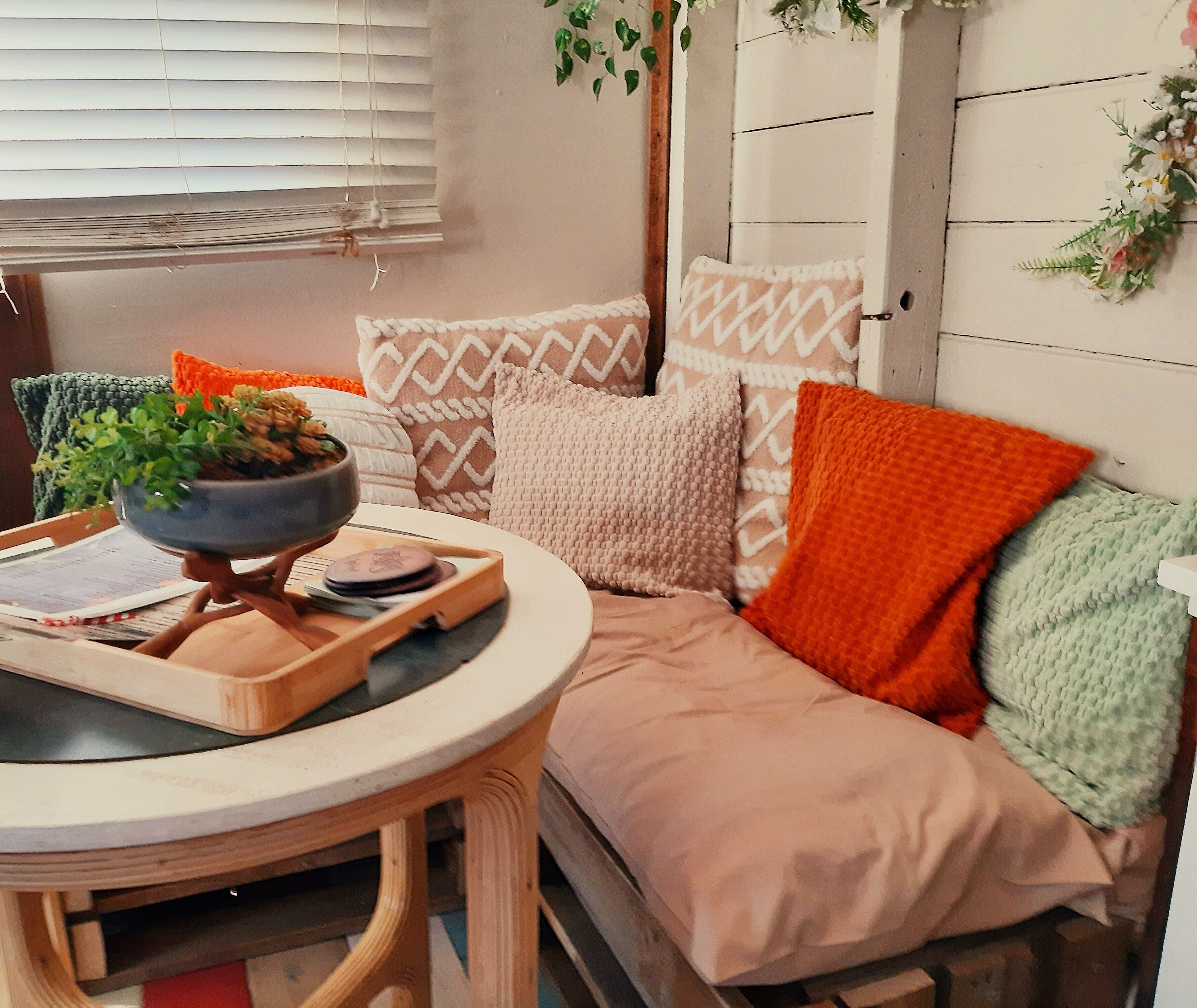 A cozy room with a round wooden table, a sofa with patterned cushions, and a window with blinds.