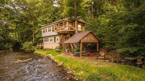 Eine Holzhütte mit einer Holzterrasse und einer kleinen Veranda, umgeben von einem Wald und einem Bach.