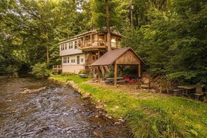 Une cabane dotée d’une terrasse en bois et d’un petit porche, entourée d’une forêt et bordée par un ruisseau.