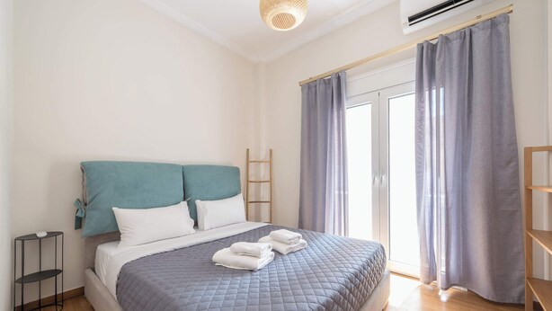 A neatly made bed with a grey quilt, a teal headboard, and a small bedside table with a lamp. - Stylish 2 Bd Urban Retreat Near Panormou Metro (Athina)