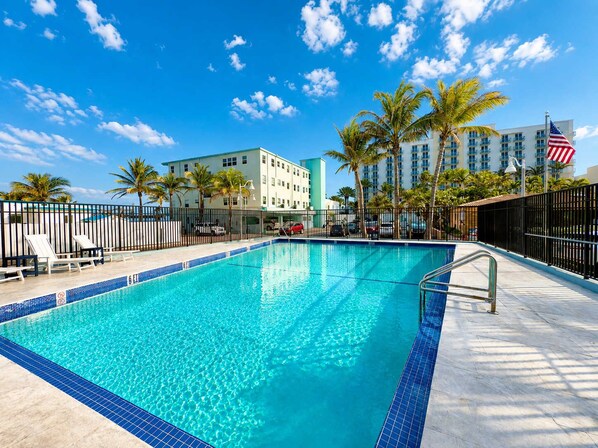 Una piscina con borde de azulejos, sillas de descanso, palmeras y un edificio de hotel al fondo.