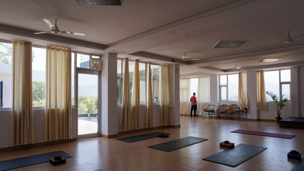 A spacious room with large windows, yoga mats, and a ceiling fan. - Yogamu India (Rishikesh)