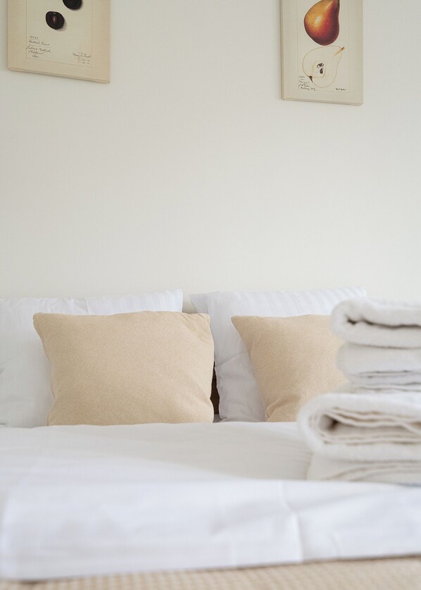 A neatly made bed with white linens and beige pillows, a folded white towel on the bed, and two framed botanical prints on the wall.
