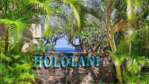 Un muro de piedra con el nombre "HOLOLANI" en letras grandes, rodeado de vegetación exuberante y vista al océano.
