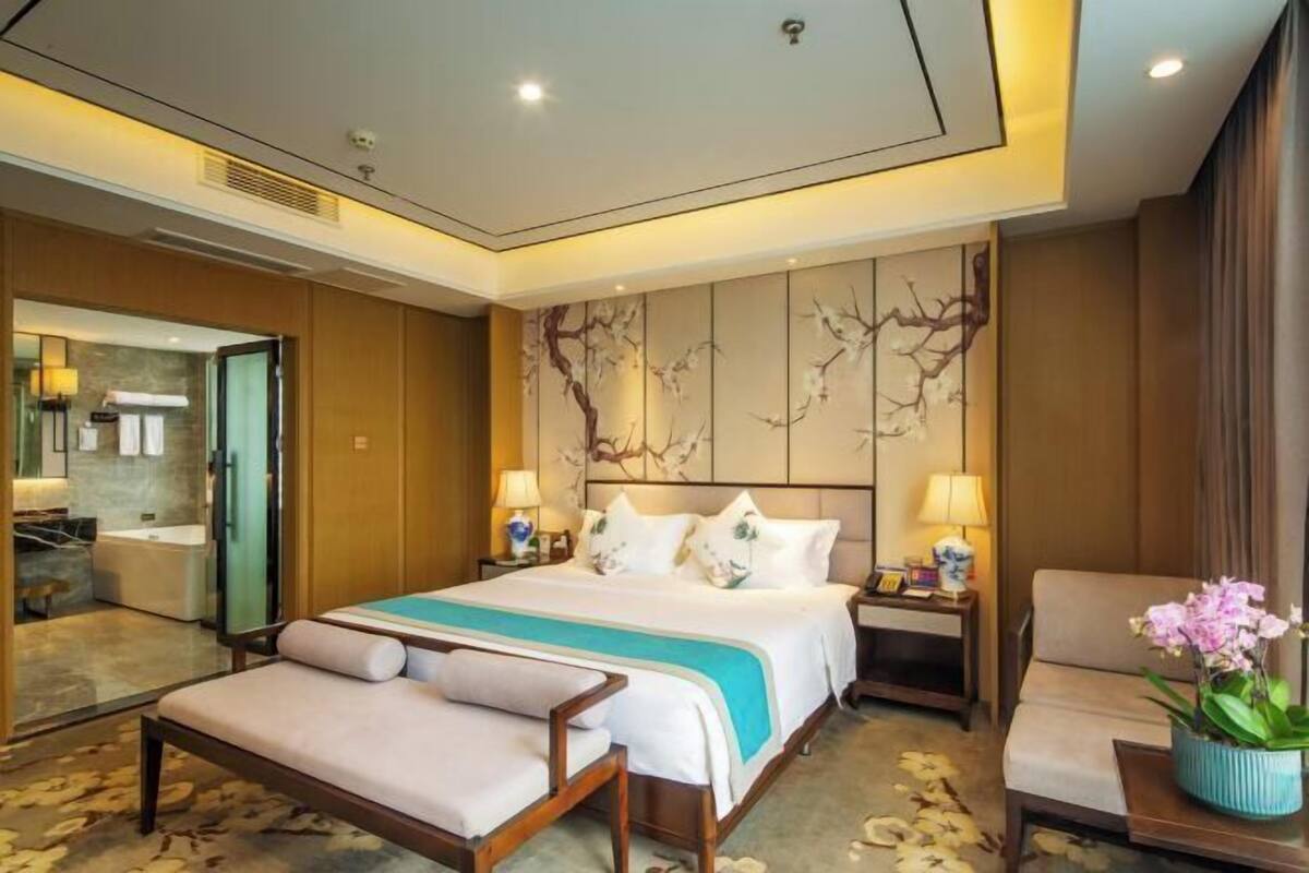 A hotel room with a bed, a sofa, a nightstand, a lamp, and a potted plant.