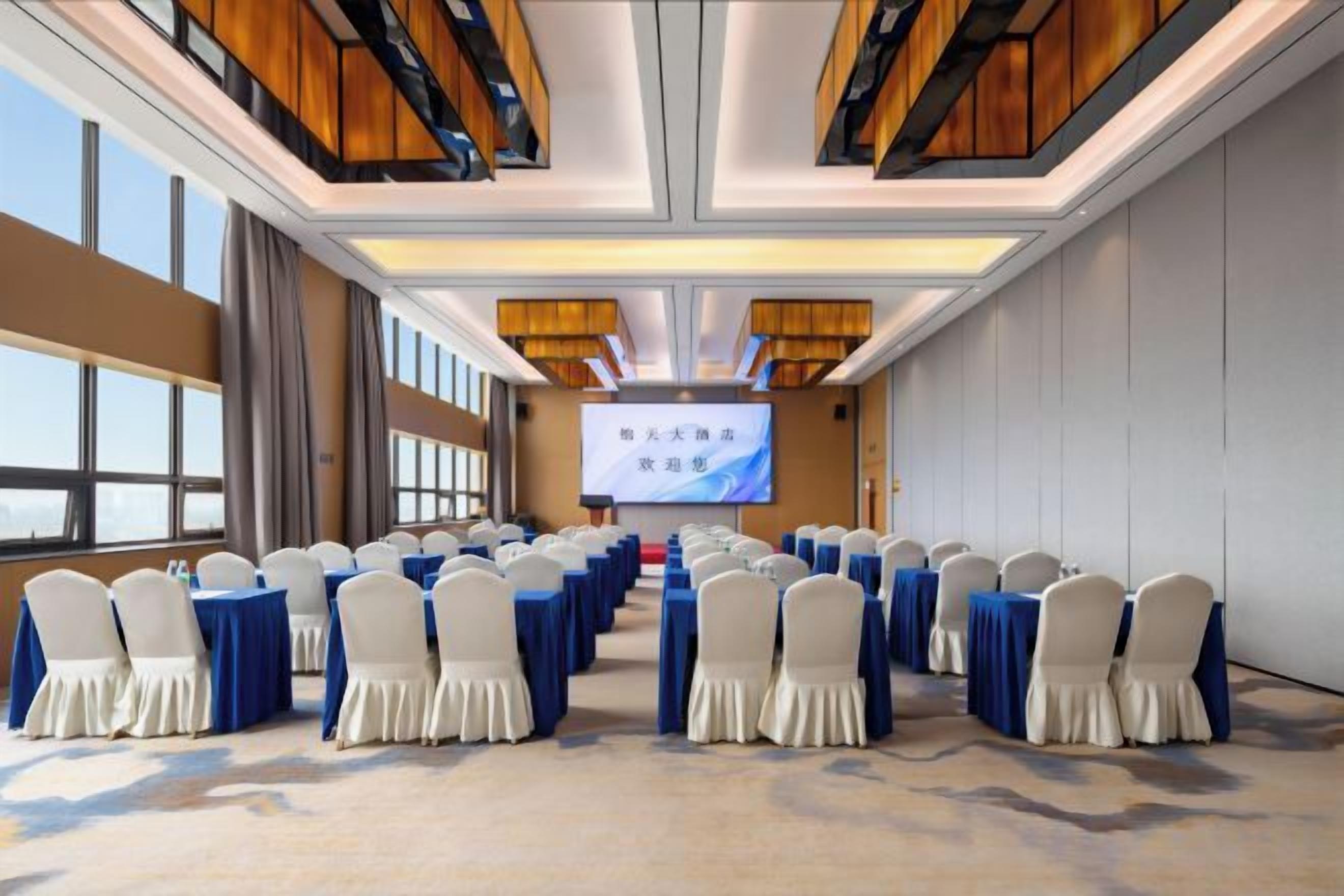 Un salón de conferencias con filas de mesas y sillas, una pantalla grande al frente y vistas del cielo a través de amplias ventanas.