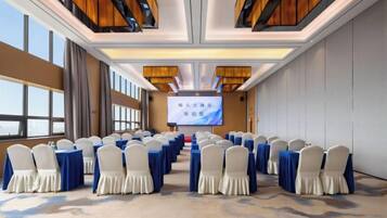 Un salón de conferencias con filas de mesas y sillas, una pantalla grande al frente y vistas del cielo a través de amplias ventanas.