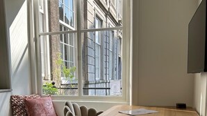 Een knus vertrek met een houten tafel en twee stoelen, een raam met uitzicht op gebouwen en een televisie aan de muur.