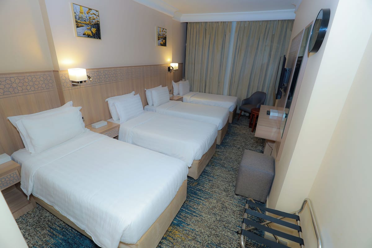 Habitación de hotel con tres camas individuales, un escritorio, una silla, una mesita, un televisor y una ventana con cortinas.