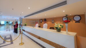 A hotel reception area with a reception desk, two staff members, a clock, and a sign indicating the location as Jakarta.