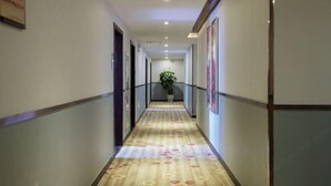 A hotel hallway with a patterned carpet, framed artwork on the walls, and a potted plant at the end. - Borui Business Hotel (Tongxiang)