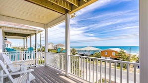 Une véranda en bois avec des balustrades blanches, un espace de détente couvert avec des chaises, et une vue sur l’océan et les maisons de plage.