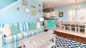 Un salon avec un canapé à rayures, un fauteuil turquoise, une table à manger avec des chaises blanches, et une cuisine en arrière-plan.