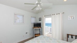 Une chambre dotée d’un ventilateur de plafond, d’un téléviseur posé sur un meuble en bois, d’un lit, d’une fenêtre avec des stores et offrant une vue sur l’océan par une porte.