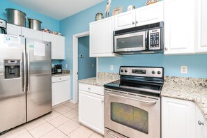 A kitchen with stainless steel appliances, including a refrigerator, microwave, and oven, with white cabinetry and a blue wall. - Barefoot Bumblebee - 2nd Row 5BR Home - Ocean Views from Decks and Porches (Surf City)