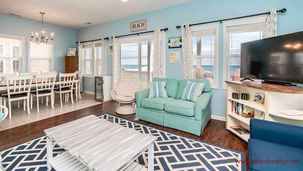 Un salon spacieux avec un canapé de couleur turquoise, un coin repas avec des chaises blanches, un téléviseur posé sur un meuble en bois et une vue sur la plage grâce à de grandes fenêtres.