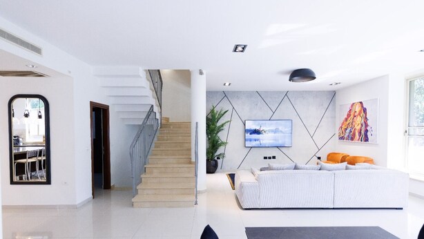 A modern living room with a sofa, a TV mounted on the wall, and a staircase leading to another room. - HaSharon Tranquil Villa by Sea N' Rent (Hod HaSharon)
