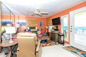 A room with a ceiling fan, a television, a sofa, a coffee table, and a view of the ocean through a door.