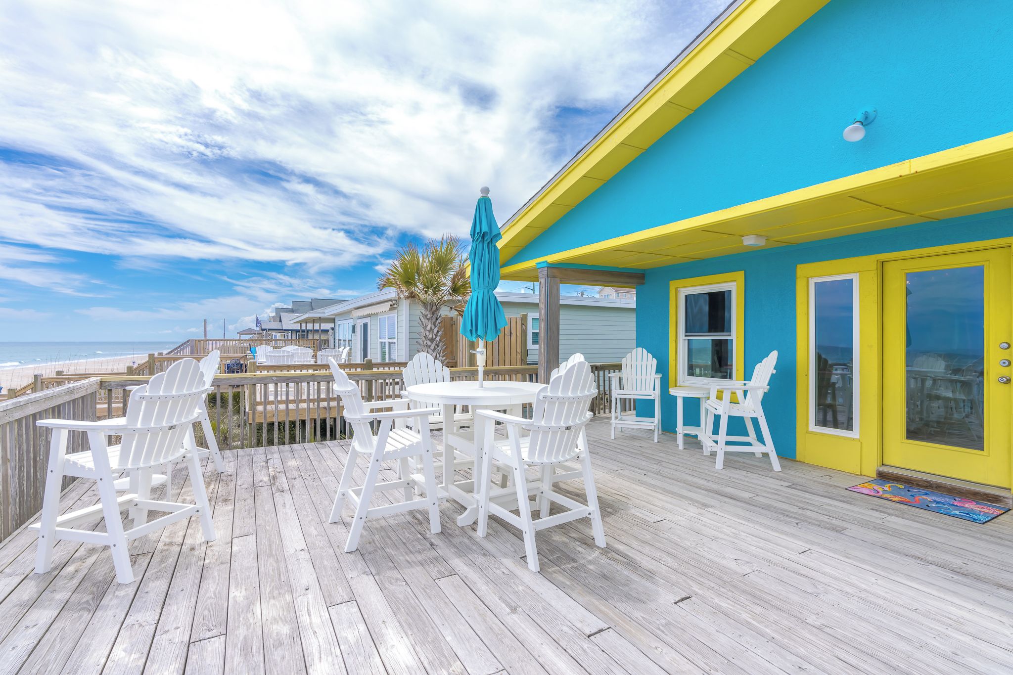 Una terraza de madera con sillas blancas y una mesa, una sombrilla azul y un edificio amarillo con ventanas.